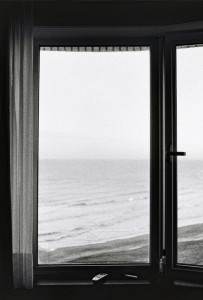 A monochrome photograph of a closed window with sheer curtains drawn to the sides. Beyond the window, the view reveals an expansive, calm ocean with gentle waves meeting the shoreline in Ostende. A remote control rests on the windowsill.